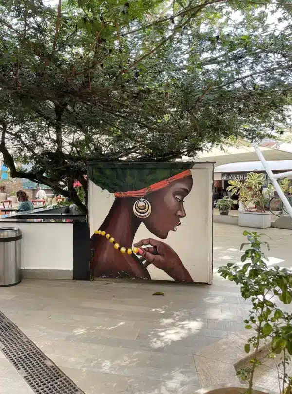 Art mural of an African woman wearing jewelry at Village Market Nairobi, surrounded by outdoor cafes and greenery.