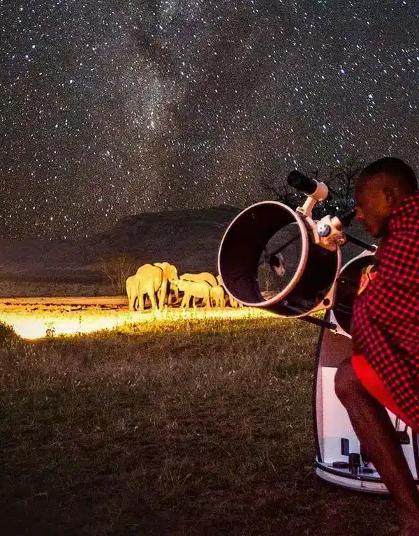 Nighttime astro-tourism in Samburu as a Samburu guide adjusts a telescope with elephants grazing beneath a star-filled sky near Samburu Sopa.